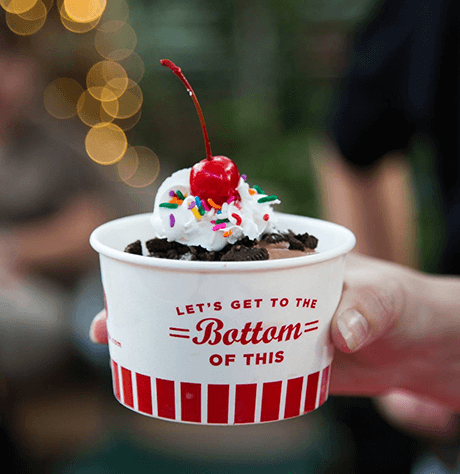 A hand holding a ice cream cup with cookies, whipped cream, sprinkles and a cherry on top.